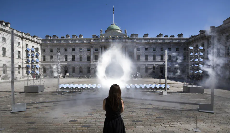 Clear sky. Outside in front of a palace-like facility, a number of circular mirrors are mounted on the floor and on towers erected on either side. In the center, a circle of light reflecting and collecting the sun’s rays floats in the fog. A single viewer stands in the center looking at it.