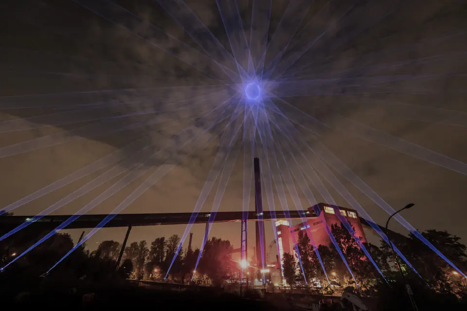 Night. On a park-like site of a former coal mine, laser beams emitted from numerous blue-violet lights on the ground are gathered overhead to form a ball of light.