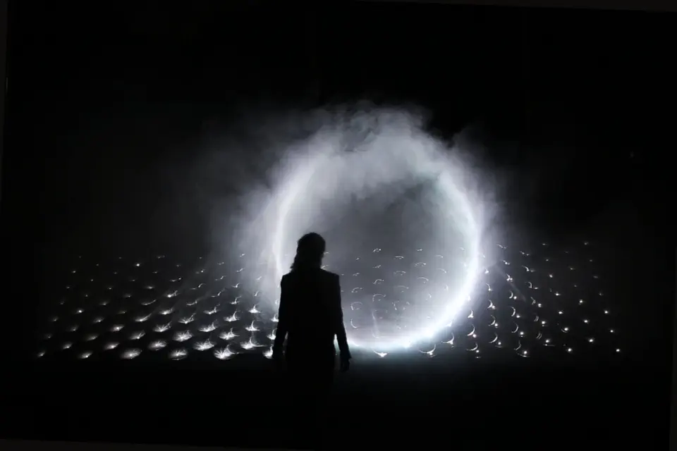 Circular mirrors lined up regularly in a dark room. A circle of light rises out of the fog. The viewer stands in front of the central circle.