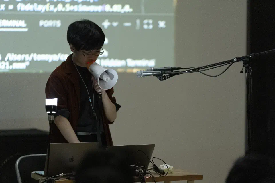 やや左側からアップで撮影された松浦のパフォーマンスの様子。左手で口の手前にメガフォンを持ってきており、右側からブームスタンドで2つのマイクロフォンがメガフォンの先を向くように設置されている。